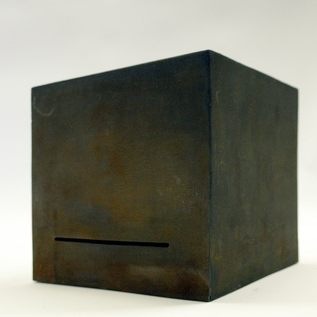 Dull green/black metal cube facing camera at a slight angle, with a single skinny, wide slit at the bottom of the front face, against a white background