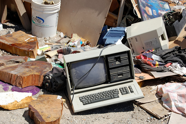 Photo of a garbage dump site with a very old personal computer with 5.25 inch diskette drives in the foreground. In the background you can see an old Apple Macintosh and other assorted garbage.